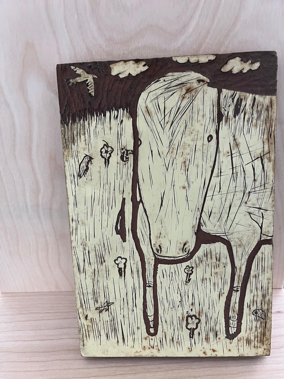 Image of a woodcut printing block with a stylized cow in a field of flowers with a bird and clouds in the sky behind it