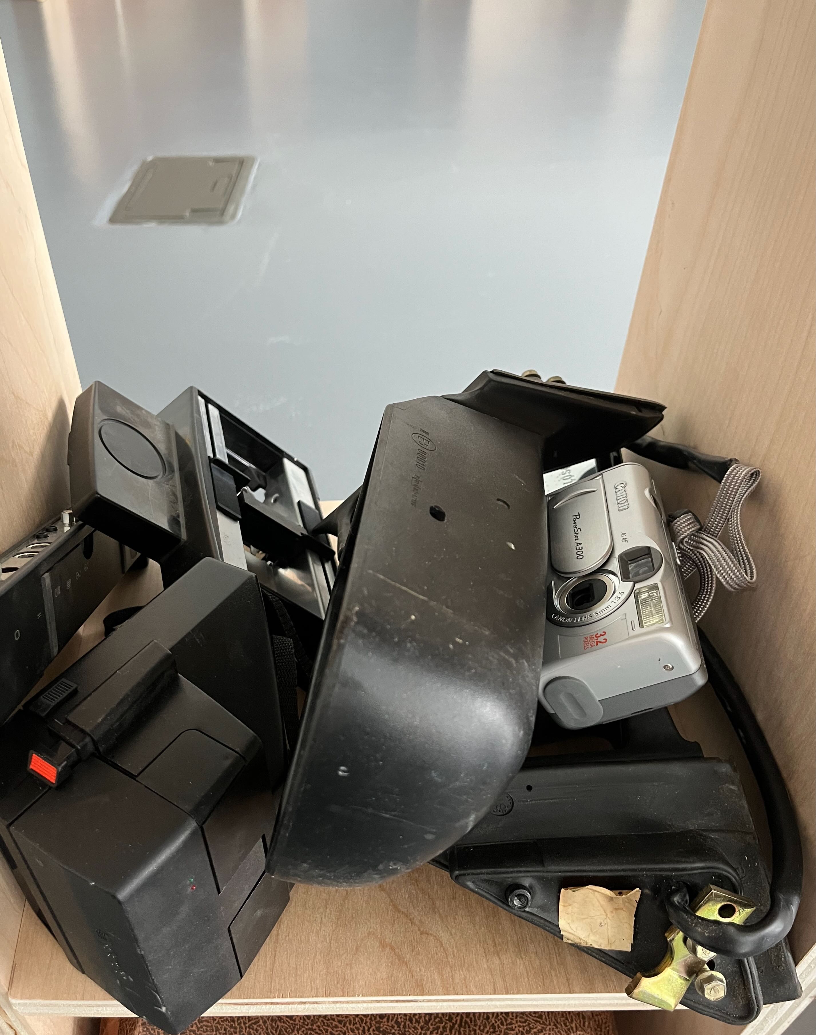 Image of a pile of objects including an old point and shoot camera, a polaroid, two car side mirrors, and some miscellaneous e-waste objects